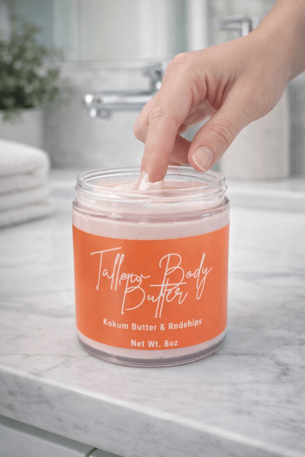 Hand dipping into a jar of Tallow Body Butter, featuring kokum butter and rosehips, on a marble countertop.